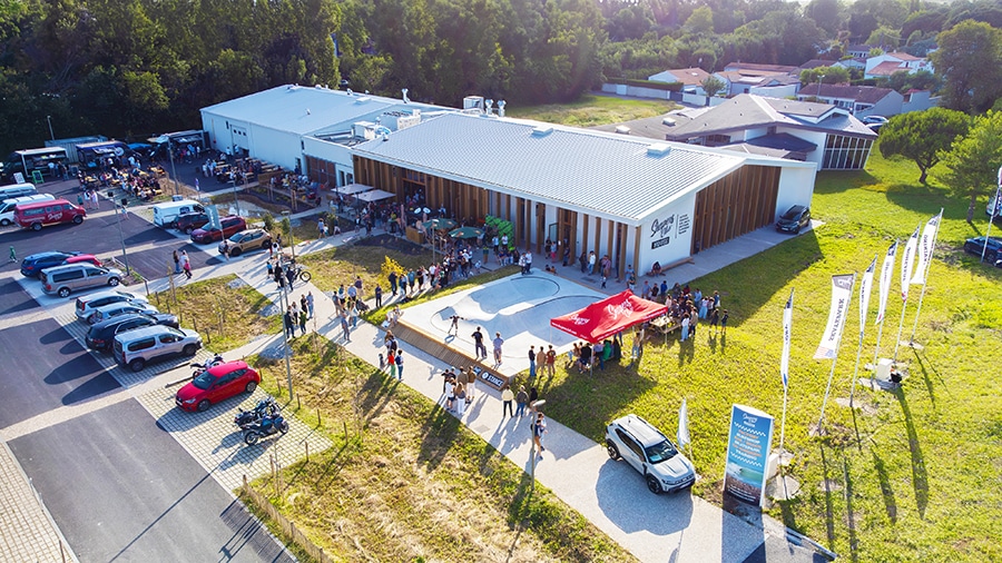 Une vue aérienne d'un événement communautaire à l'extérieur d'un bâtiment moderne révèle une scène accueillante avec un grand rassemblement de personnes, des tentes, des voitures garées et de la verdure environnante.