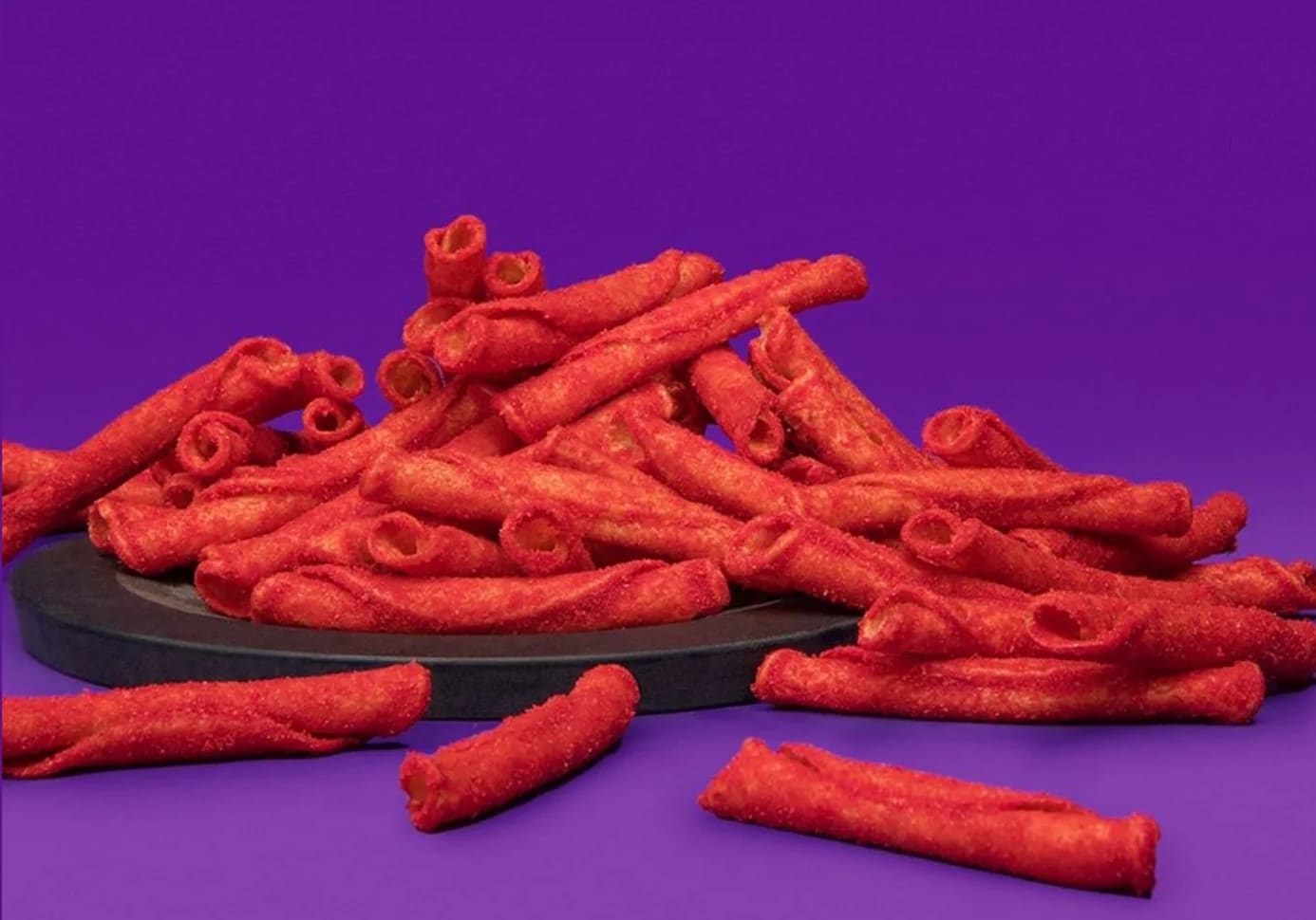 Des snacks de maïs rouge roulés et croquants sont empilés sur une surface noire sur un fond violet, évoquant l'aura vibrante d'une véritable Révolution Épicée.