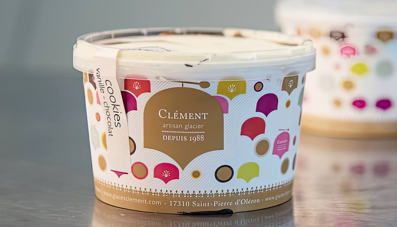 Un pot de crème glacée Clément délices glacés avec un motif à pois colorés sur l'étiquette est posé sur une surface réfléchissante. La saveur indiquée est biscuits et chocolat vanille. Le nom de la marque et l'adresse sont bien visibles, faisant allusion à sa qualité supérieure disponible au Shapers Club House.