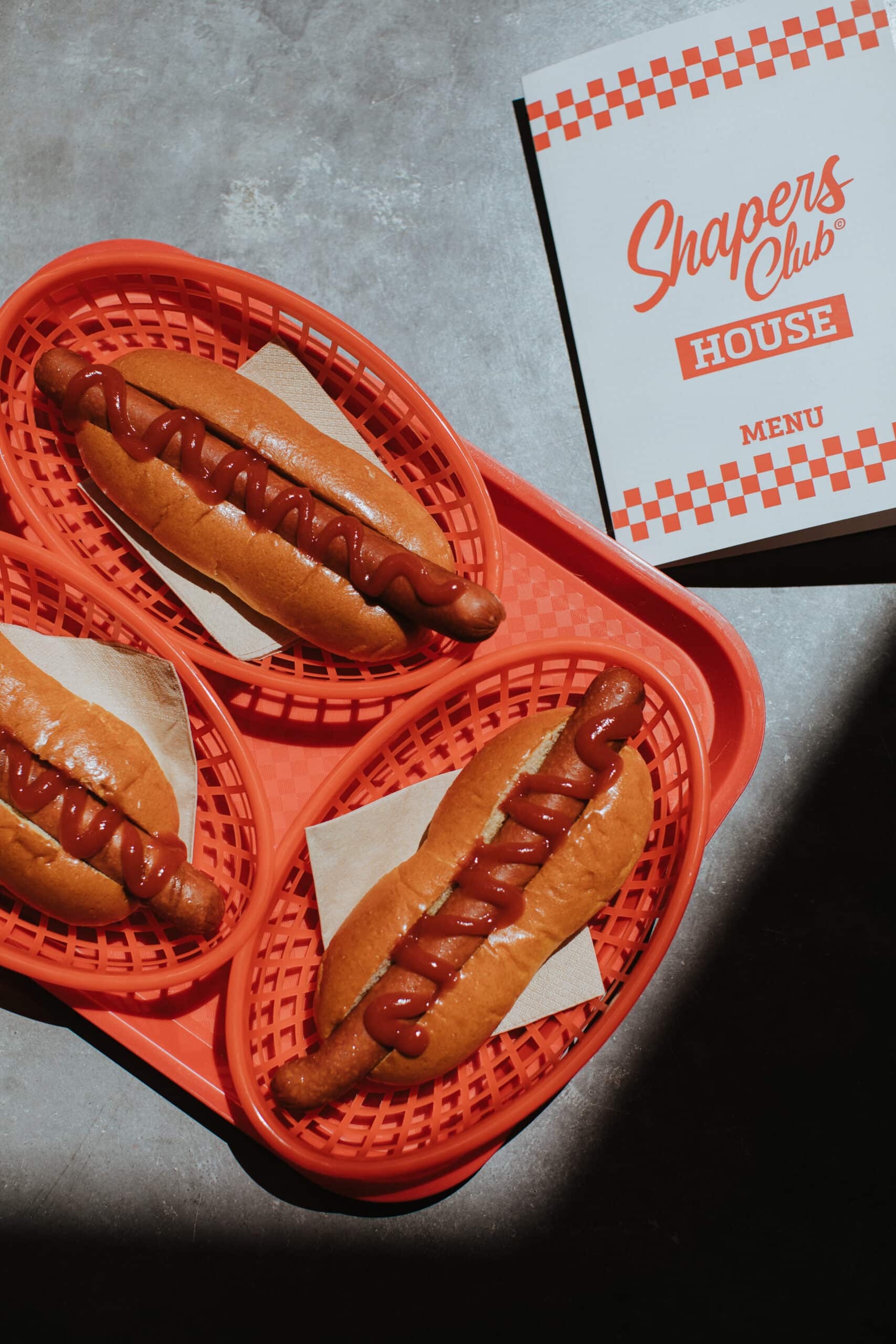 Trois hot-dogs avec du ketchup dans des paniers rouges sur un plateau rouge, à côté d'un menu intitulé « Shapers Club House », créent un accueil chaleureux pour les invités.