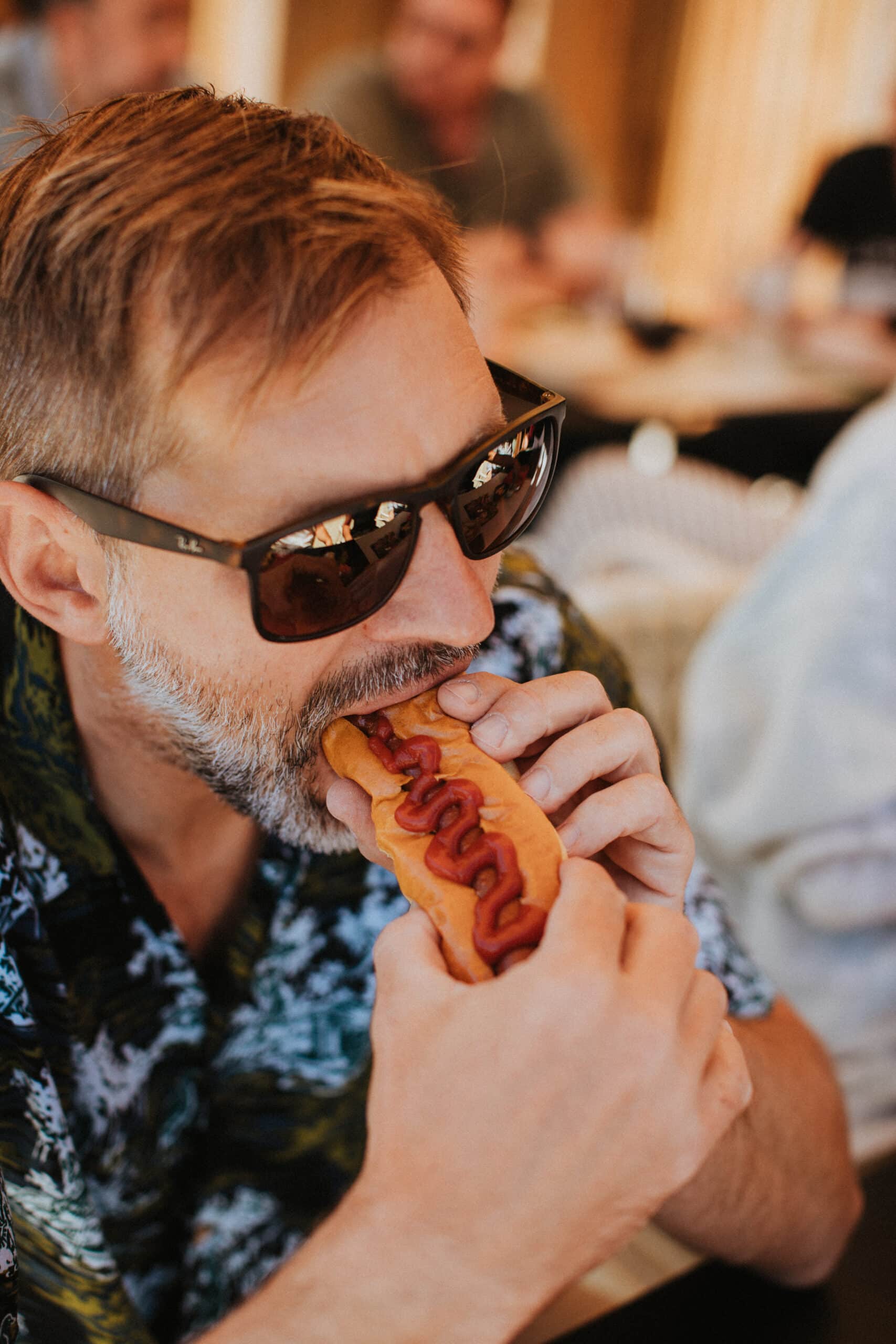 Un homme portant des lunettes de soleil et une chemise à motifs savoure l'accueil d'un hot-dog garni de ketchup.