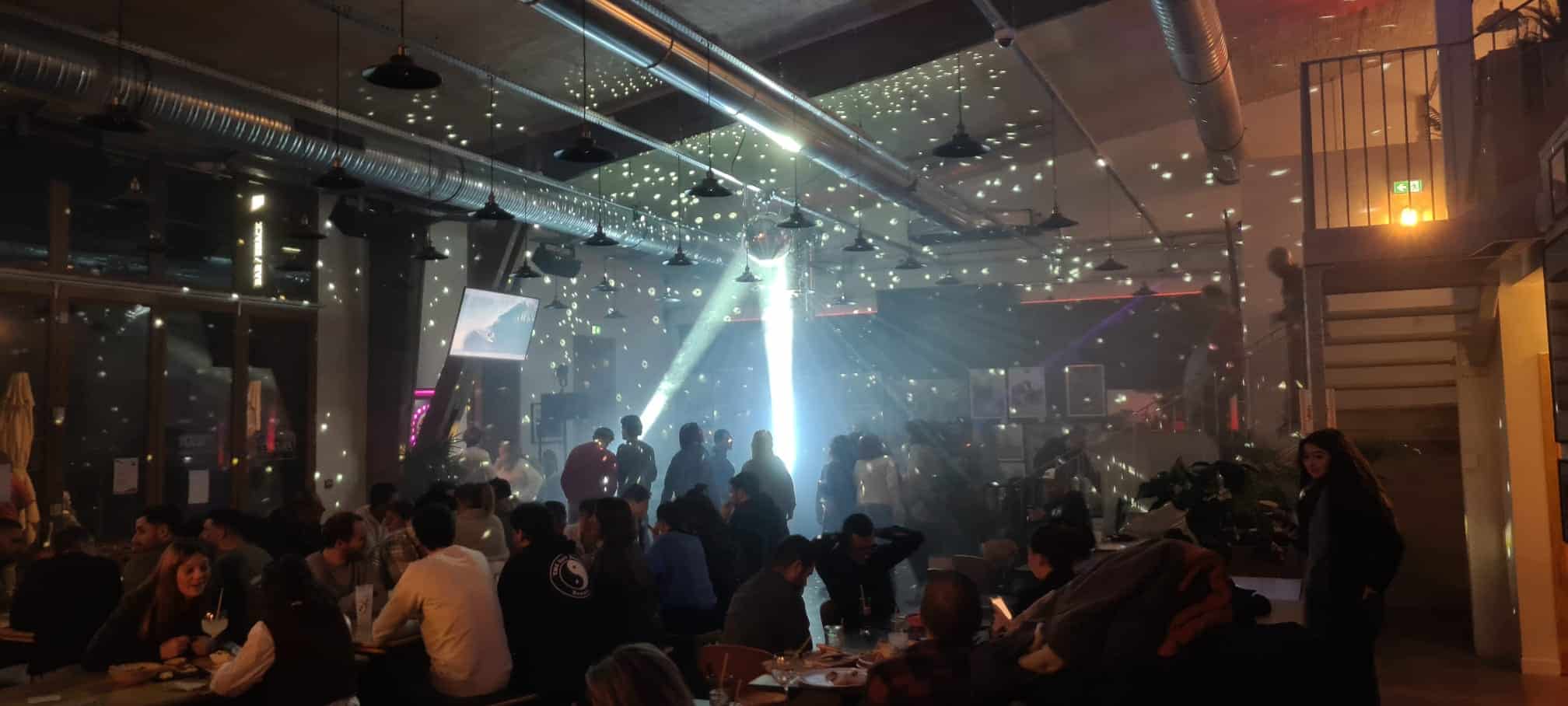 Une salle intérieure à la lumière tamisée accueille une soirée animée avec des groupes de personnes assises et debout. Des faisceaux lumineux et des reflets de boules à facettes créent des motifs surprenants sur les murs et le plafond, ajoutant une touche de magie à cette soirée de février.